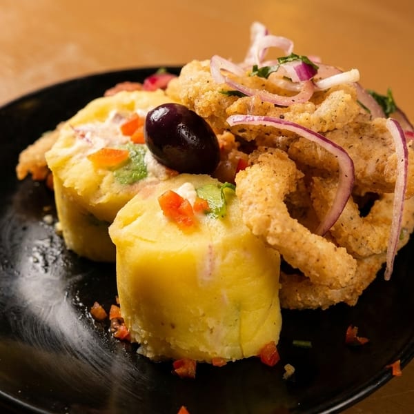 Peruvian causa rellena with fried calamari, red onion, and a black olive on a black plate.