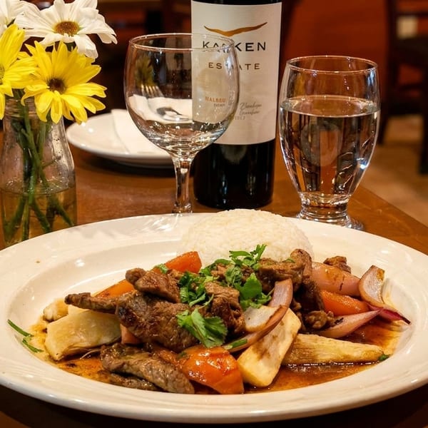 A plate of Lomo Saltado with rice, wine, water, and flowers on a wooden table.