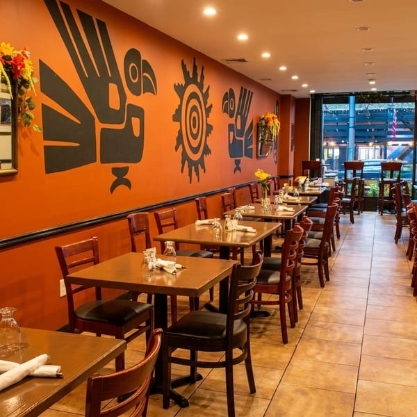 Restaurant interior with orange walls, tribal art, wooden tables, chairs, and flowers.