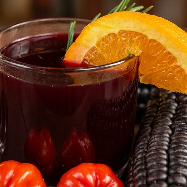 Purple corn drink in a glass mug, garnished with orange and rosemary, surrounded by purple corn and peppers.